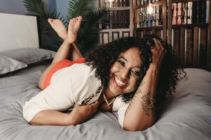A woman with curly hair lying on a bed, smiling confidently in a boudoir photoshoot.