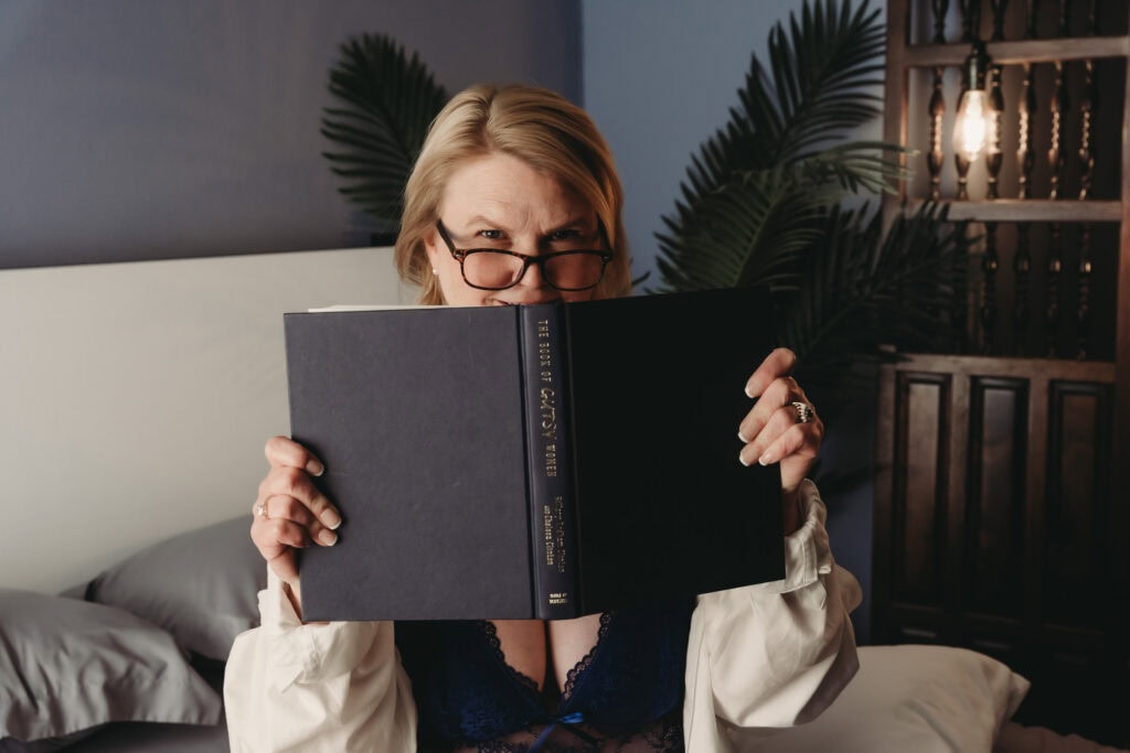 A woman with glasses holding a book in front of her face in a cozy, stylish bedroom.