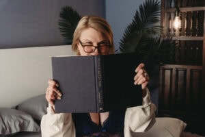 A woman with glasses holding a book in front of her face in a cozy, stylish bedroom.